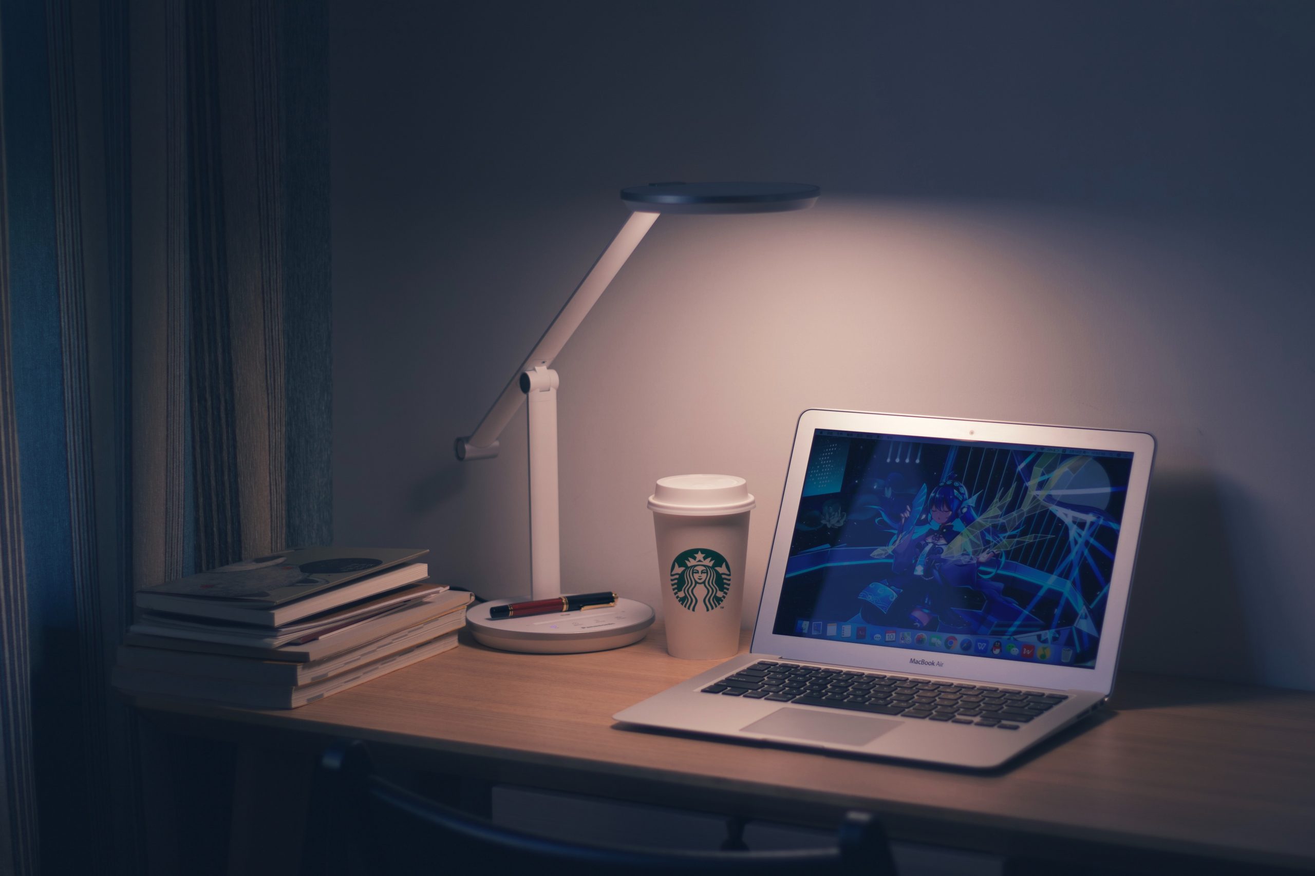 A hazy desk light shines on a wooden desk with a laptop and Starbucks coffee cup.